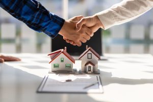 Two people shaking hands over two small house figurines and documents